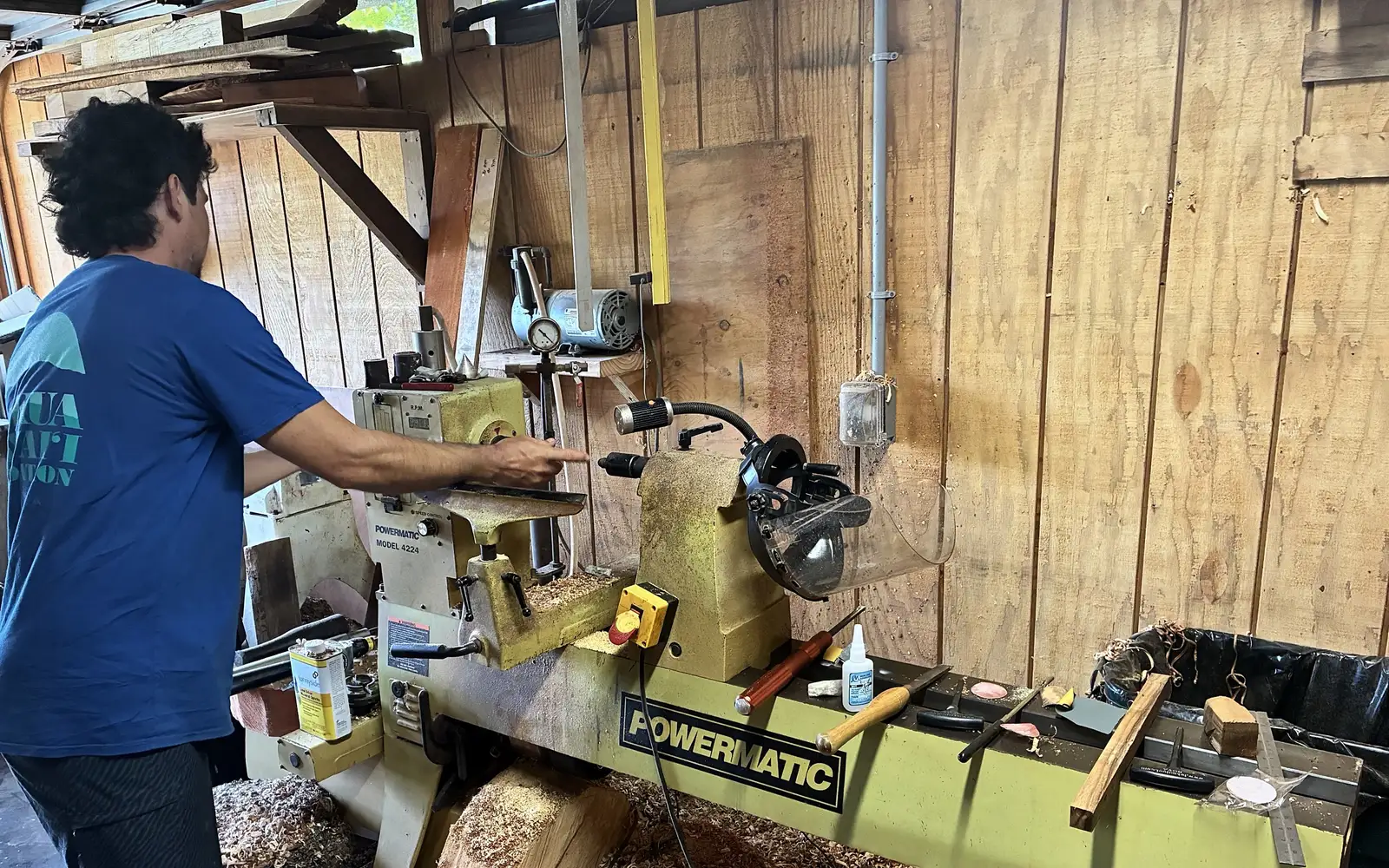 Kaleb at his Powermatic lathe, shavings heaped on the shop floor beneath it