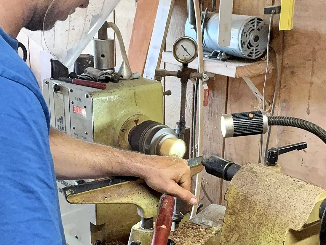 Kaleb at the lathe in his face shield, both hands on the tool as the wood spins
