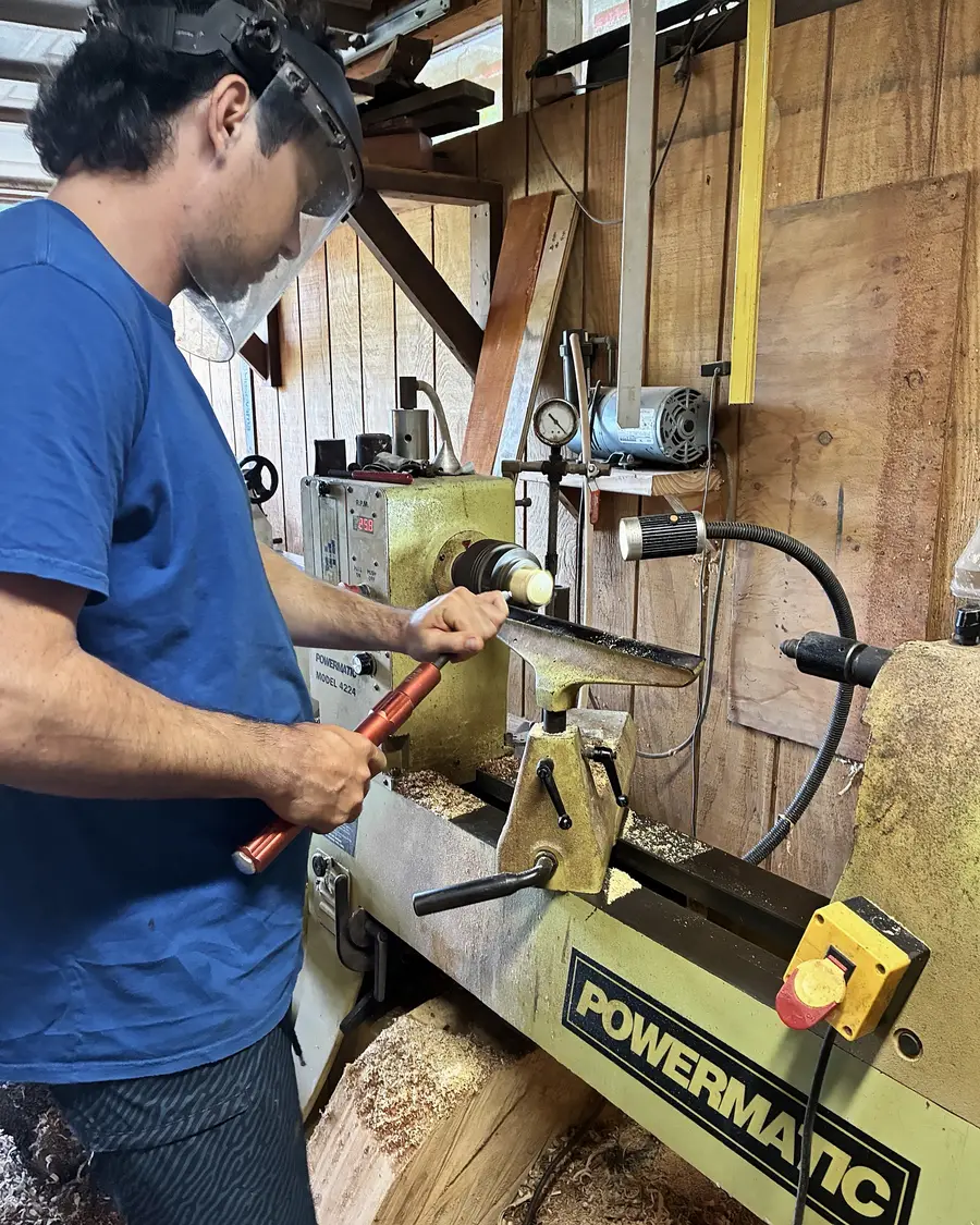 Kaleb turning at the lathe in a face shield, the tool steady on the rest and shavings on the bed